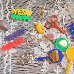 May include: Assortment of colorful plastic toys, including a toy train, a watering can, a frying pan, a mug, a pair of scissors, a wrench, and several word charms. The charms spell out "YES HAPPY", "WHAT", and "HELLO".