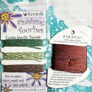 May include: A close-up of a package of Kreinik Finishing Touches Fusible Iron-On Threads. The package shows four spools of thread in green, brown, and gold. The text "Use on paper, fabric, wood, and even glass!" is printed on the package. A separate package of Fiesta 6-Ply Rayon Floss in a rust color is also shown. The text "FIESTA! 6-Ply Rayon Floss For Needlepoint, Cross Stitch, Embroidery" is printed on the package.