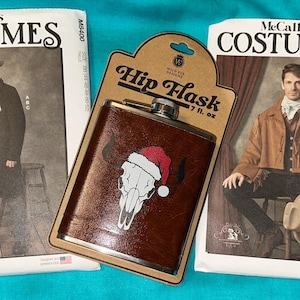 May include: A brown leather hip flask with a skull wearing a Santa hat, next to two McCall's costume patterns. The flask holds 7 fl oz. The costume patterns feature men in Western-style outfits, including coats, hats, and vests.