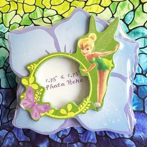 May include: A purple and green wooden picture frame with a Tinkerbell character and a butterfly design. The frame has a 1.75" x 1.75" opening with the text "Photo Here".