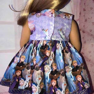 K&ouml;nnte beinhalten: &Auml;rmelloses Puppenkleid mit einem lebendigen Aufdruck von Figuren aus dem Animationsfilm "Die Eisk&ouml;nigin". Das Kleid hat ein lila Oberteil mit Schneeflocken- und Olaf-Designs. Der Rock zeigt Elsa, Anna, Kristoff und Olaf. Das Kleid wird mit Kn&ouml;pfen geschlossen.