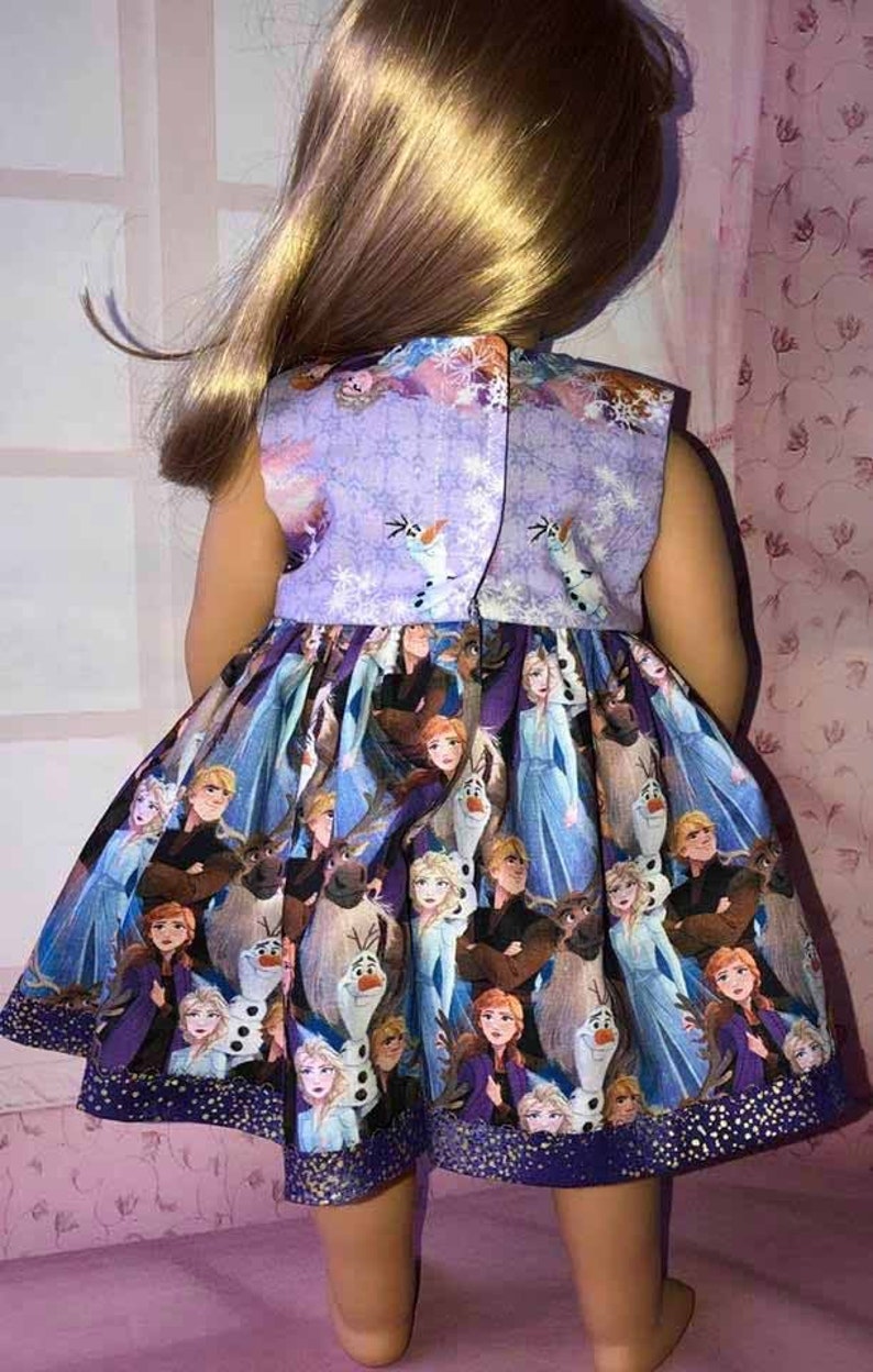 K&ouml;nnte beinhalten: &Auml;rmelloses Puppenkleid mit einem lebendigen Aufdruck von Figuren aus dem Animationsfilm "Die Eisk&ouml;nigin". Das Kleid hat ein lila Oberteil mit Schneeflocken- und Olaf-Designs. Der Rock zeigt Elsa, Anna, Kristoff und Olaf. Das Kleid wird mit Kn&ouml;pfen geschlossen.
