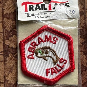 May include: A red and white embroidered patch with the text "Adams Falls" and a brown fish with green eyes. The patch is packaged in a clear plastic bag with a white card that reads "Trail Tags, LYMA enterprises, 1972, P.O. Box 5058."