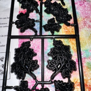May include: Black plastic sprue with various floral and leaf shapes. The sprue is designed for cutting out individual pieces. The background is a colorful, patterned fabric. Text on the left side reads "ON FRO".