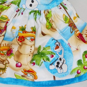 Pode incluir: Um tecido branco com uma bainha azul apresenta uma cena de praia tropical com bonecos de neve usando &oacute;culos de sol e chap&eacute;us, palmeiras e Olaf de Frozen.