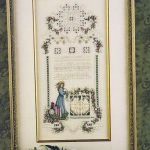 May include: Framed cross-stitch artwork featuring a woman in a blue dress watering flowers by a window. The design includes floral and geometric patterns, with a gold frame. The text "Cross 'N Patch" is visible.