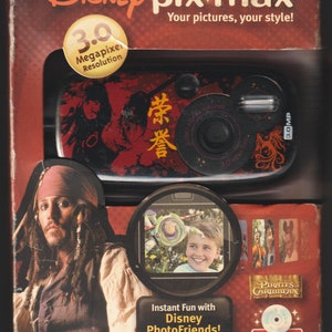 May include: A red, black, and gold Disney Pix-Max 3.0 Megapixel camera with a Pirates of the Caribbean theme. The camera features a picture of Captain Jack Sparrow on the front. The box also includes a picture of a child taking a picture with a toy Buzz Lightyear.
