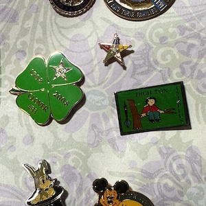 May include: A collection of colorful enamel pins, including a green four-leaf clover pin with "1908 Clover 2008" text, a Mickey Mouse birthday pin, and a "Hat Trick" pin. Other pins feature text and star designs.