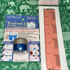 May include: A Thread Magic thread conditioner in a blue and silver container on a blue backing card, next to a peach-colored ruler on a white backing card. The ruler shows measurements in inches, with the scale clearly visible up to 6 inches.