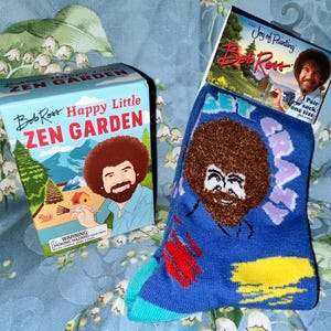 May include: A box for a "Happy Little Zen Garden" and a pair of blue crew socks, both featuring Bob Ross's image. The socks have a colorful design with paint strokes and the word "CREATE". The box has a landscape illustration.