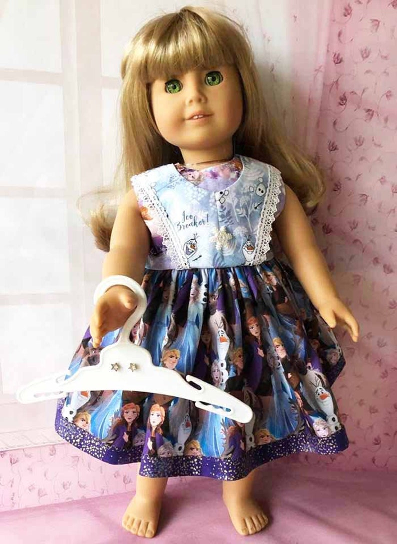 K&ouml;nnte beinhalten: Puppe in einem ma&szlig;geschneiderten Kleid mit Figuren aus dem Animationsfilm. Das Kleid hat ein blaues Oberteil mit dem Text "Ice Breaker!" und einen lila Rock mit Charakterillustrationen. Die Puppe h&auml;lt einen wei&szlig;en Kleiderb&uuml;gel.