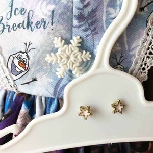 K&ouml;nnte beinhalten: Ein wei&szlig;er Kunststoffb&uuml;gel pr&auml;sentiert ein hellblaues Kleid mit einem Schneeflocken- und sternf&ouml;rmigen Ohrringen. Das Kleid zeigt den Text "Ice Breaker!" und eine Cartoonfigur. Die Ohrringe sind goldfarben mit einem rosa Mittelteil.