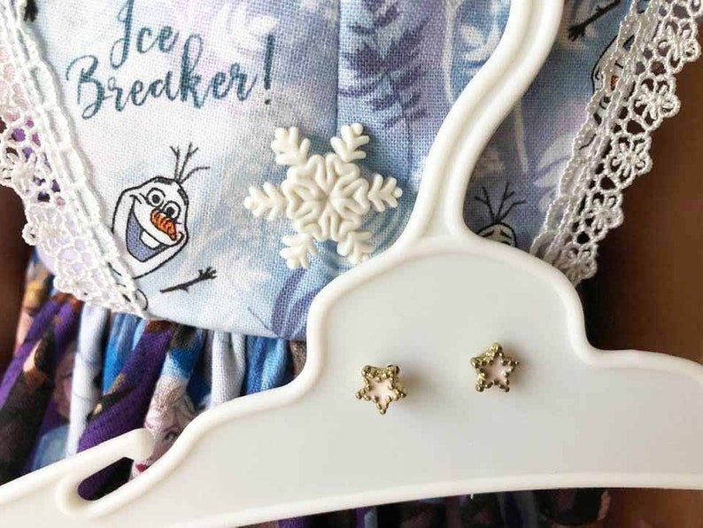 K&ouml;nnte beinhalten: Ein wei&szlig;er Kunststoffb&uuml;gel pr&auml;sentiert ein hellblaues Kleid mit einem Schneeflocken- und sternf&ouml;rmigen Ohrringen. Das Kleid zeigt den Text "Ice Breaker!" und eine Cartoonfigur. Die Ohrringe sind goldfarben mit einem rosa Mittelteil.
