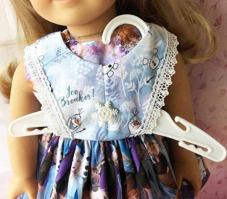 K&ouml;nnte beinhalten: Hellblaues Puppenkleid mit wei&szlig;er Spitzenborte und Schneeflockenverzierung. Das Kleid zeigt einen Aufdruck von Figuren und den Text "Ice Breaker!". Es h&auml;ngt an einem wei&szlig;en Kleiderb&uuml;gel.