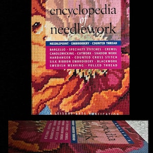 May include: A hardcover book titled "Encyclopedia of Needlework" by Donna Kooler. The cover features a colorful needlepoint design in shades of orange, red, and pink. The book's spine and pages are visible, with a list of needlework techniques.