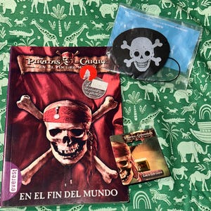 May include: A Pirates of the Caribbean En El Fin Del Mundo book featuring a skull and crossbones graphic, a black eye patch with a white skull and crossbones design in a plastic bag, and a small Pirates of the Caribbean Dead Man's Chest charm.
