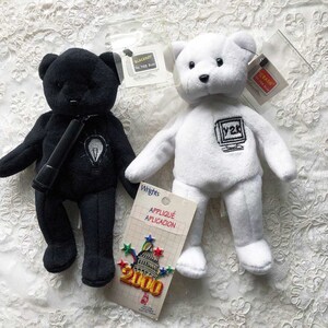 Puede incluir: Dos osos de peluche, uno negro y otro blanco, con detalles bordados relacionados con el año 2000. El oso negro tiene una bombilla y un marcador, mientras que el oso blanco tiene una pantalla de ordenador con "Y2K" en ella. Ambos osos tienen pequeñas etiquetas con texto relacionado con el año 2000.