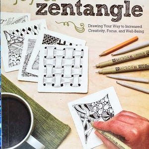 May include: A book titled "Joy of Zentangle" with a drawing of a hand using a Micron pen to create a Zentangle pattern. The book cover features the text "Drawing Your Way to Increased Creativity, Focus, and Well-Being".