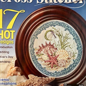 May include: The June 2010 issue of The Cross Stitcher magazine features a circular wooden frame containing a cross-stitch pattern of a starfish, seashells, and seaweed. A large, textured starfish rests in the foreground, with additional shells.