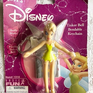 May include: A bendable Tinker Bell keychain with a green dress and silver shoes. The keychain is packaged on a pink and purple background with the words "Disney", "Tinker Bell", "Bendable Keychain", and "Really Works!" printed on it.