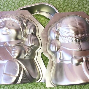 May include: Two silver metal cake pans shaped like a doll. The doll on the left is sitting with a present in her lap. The doll on the right is standing with a bow on her dress.