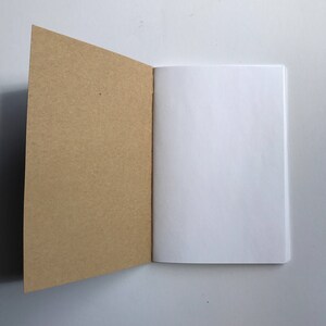 May include: An open notebook with a kraft paper cover and blank white pages. The left cover is angled open, revealing the interior pages. The notebook is set against a white background, creating a clean and simple aesthetic.