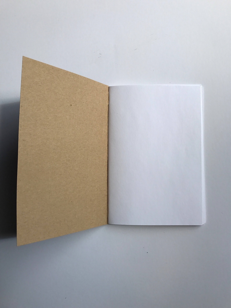 May include: An open notebook with a kraft paper cover and blank white pages. The left cover is angled open, revealing the interior pages. The notebook is set against a white background, creating a clean and simple aesthetic.