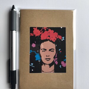 May include: A greeting card featuring a portrait of a woman with a floral headdress. The portrait is in shades of pink and black, with red, blue, and purple floral accents. The card is in a clear plastic sleeve, with a pen to the left.