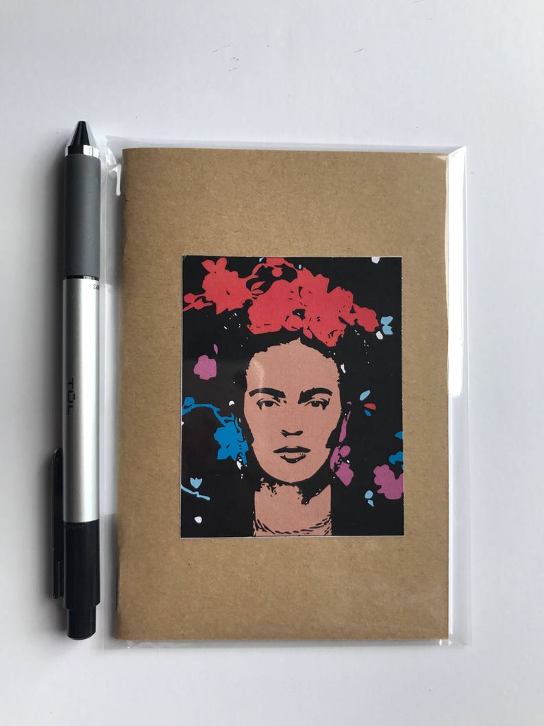 May include: A greeting card featuring a portrait of a woman with a floral headdress. The portrait is in shades of pink and black, with red, blue, and purple floral accents. The card is in a clear plastic sleeve, with a pen to the left.