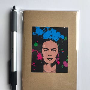 May include: A greeting card featuring a pop art style portrait of Frida Kahlo with blue and pink floral accents on a black background. The card is in a clear plastic sleeve, next to a silver and black pen.