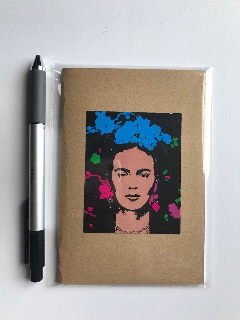 May include: A greeting card featuring a pop art style portrait of Frida Kahlo with blue and pink floral accents on a black background. The card is in a clear plastic sleeve, next to a silver and black pen.