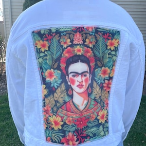 May include: White denim jacket with a colourful Frida Kahlo portrait on the back. The artwork is set against a backdrop of flowers and leaves. The jacket has a classic design with a button-up front and pockets.