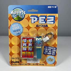 May include: A Jim Henson's Muppets PEZ keychain in original packaging. The set includes a blue Gonzo character dispenser and two packs of fruit candy. The card features orange circles, the PEZ logo, and text including "Ages 4 & Up" and "Total Candy Net Wt. .58 oz."
