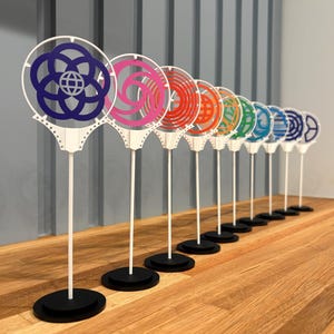 Puede incluir: Una colección de diez diseños circulares decorativos y coloridos sobre tallos blancos con bases negras. Los diseños presentan varios patrones y colores, dispuestos en fila sobre una superficie de madera.