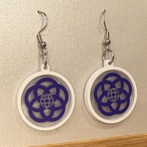 May include: A pair of white and blue EPCOT Center earrings. Each earring features a circular design with a blue floral pattern and a globe in the center. The earrings are displayed on a gray surface.