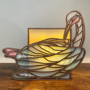 May include: A wooden bird-shaped candle holder with a layered design. The holder is made of brown wood and features a light blue, yellow, and pink color scheme. The bird is in a resting position with its head tucked under its wing.