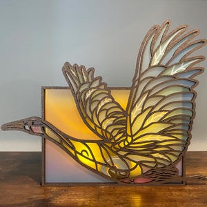 May include: A wooden bird sculpture with a stained glass design. The bird is in flight with its wings spread wide. The sculpture is illuminated from behind, creating a warm glow.