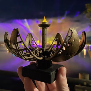 May include: A small, detailed replica of the Singapore National Stadium, featuring a golden, open-work design with a central tower and a flame on top. The replica is set on a black base.