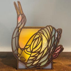 May include: A wooden candle holder shaped like a crane with a stained glass design. The crane is in a curled position with its head tucked in. The candle holder is lit and the light shines through the stained glass.