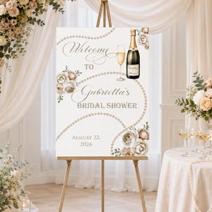 May include: A white rectangular sign on a gold easel featuring the text Welcome to Gabriella's BRIDAL SHOWER August 22, 2026. The design includes a champagne bottle, a glass of champagne, strings of pearls, and clusters of beige and white flowers.