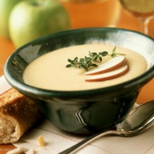 Peut inclure: Un bol de soupe crémeuse garnie de tranches de pomme et d'herbes. Une tranche de pain grillé est posée sur une serviette à côté du bol. Des pommes vertes et un verre de vin sont à l'arrière-plan. Une cuillère repose sur la serviette.