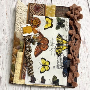 May include: A collection of fabric scraps, trim, and embellishments. Includes a butterfly print fabric, various patterned fabrics, lace, buttons, beads, and a spool of yellow thread. Brown pom-pom trim and decorative ribbon are also present.
