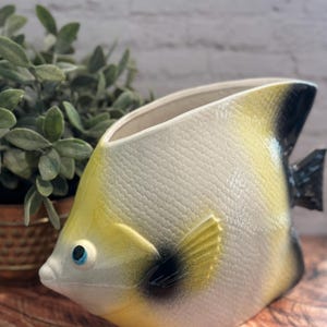May include: A ceramic fish-shaped planter or vessel featuring a textured, scale-like surface. The body is primarily cream-colored with yellow gradients, black accents on the fins and tail, and a single blue eye. The top is open for storage.