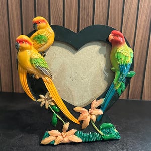 May include: A heart-shaped picture frame in black featuring three colorful parrots perched on the edges. Two yellow and green parrots are on the left side, while one red, green, and blue parrot is on the right. Peach-colored flowers and green leaves decorate the base.