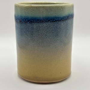 May include: A handmade ceramic tumbler with a gradient design. The top rim is a muted green, transitioning to a deep blue band, then fading into a speckled gray and finally a warm beige at the base. The cylindrical shape is simple and elegant.
