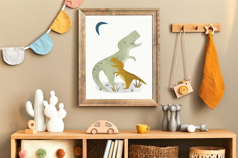 May include: A framed print of a green dinosaur with a smaller yellow dinosaur in its arms. The print has a white background and a crescent moon in the top left corner. The frame is brown wood.