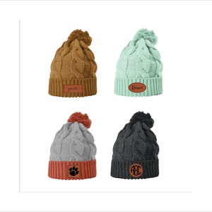 May include: Four knitted winter hats with pom-poms. The hats are in brown, light green, gray, and black. Each hat has a different leather patch with a personalized inscription: "pamola", "Blessed", a paw print, and "ABC".
