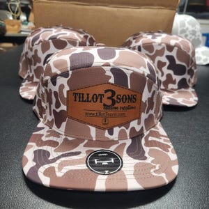 May include: A brown and white camouflage patterned baseball cap with a brown leather patch that says "Tillot 3 Sons Custom Creations www.tillot3sons.com". The cap has a brown leather patch with a drone logo and the word "Osprey" on the front.