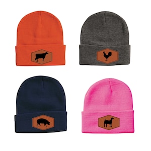 May include: Four knit beanies in different colors with a brown leather patch featuring a silhouette of a farm animal. The beanies are orange, gray, navy blue, and pink. The animals are a cow, rooster, pig, and goat.
