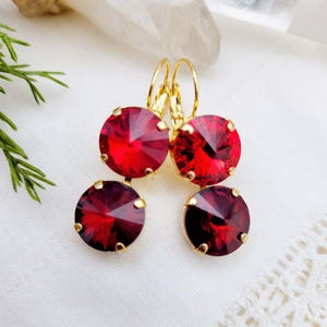 May include: A pair of gold dangle earrings with red crystal stones. The earrings have a simple design with a round crystal stone at the top and a smaller round crystal stone hanging below it.